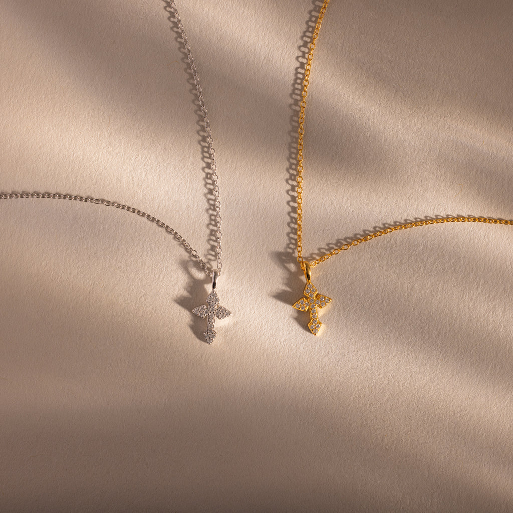 The Vintage Cross Necklace, available in silver and gold, is displayed on a softly lit beige surface with gentle shadows, highlighting its timeless style and subtle minimalist design.