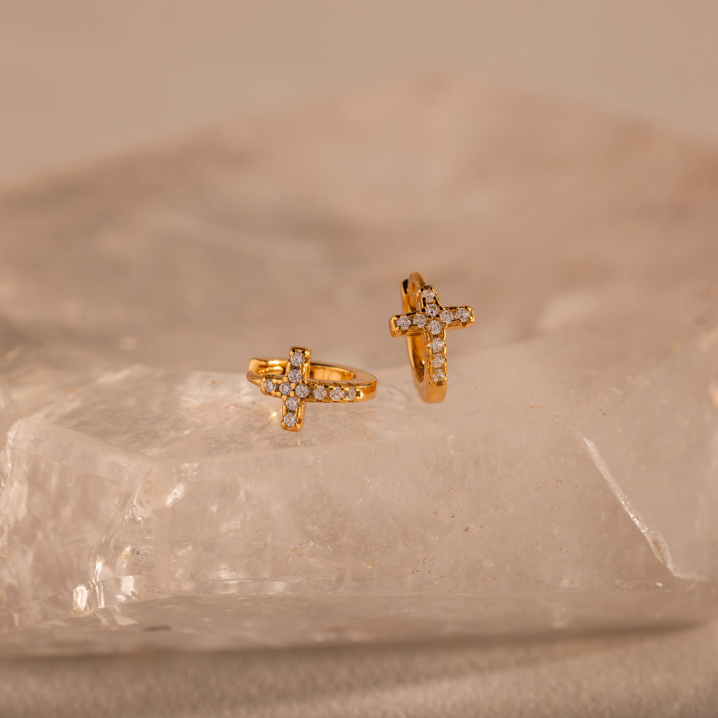Two Diamond Cross Huggies featuring gold and pavé diamonds rest on a translucent, textured crystal slab.