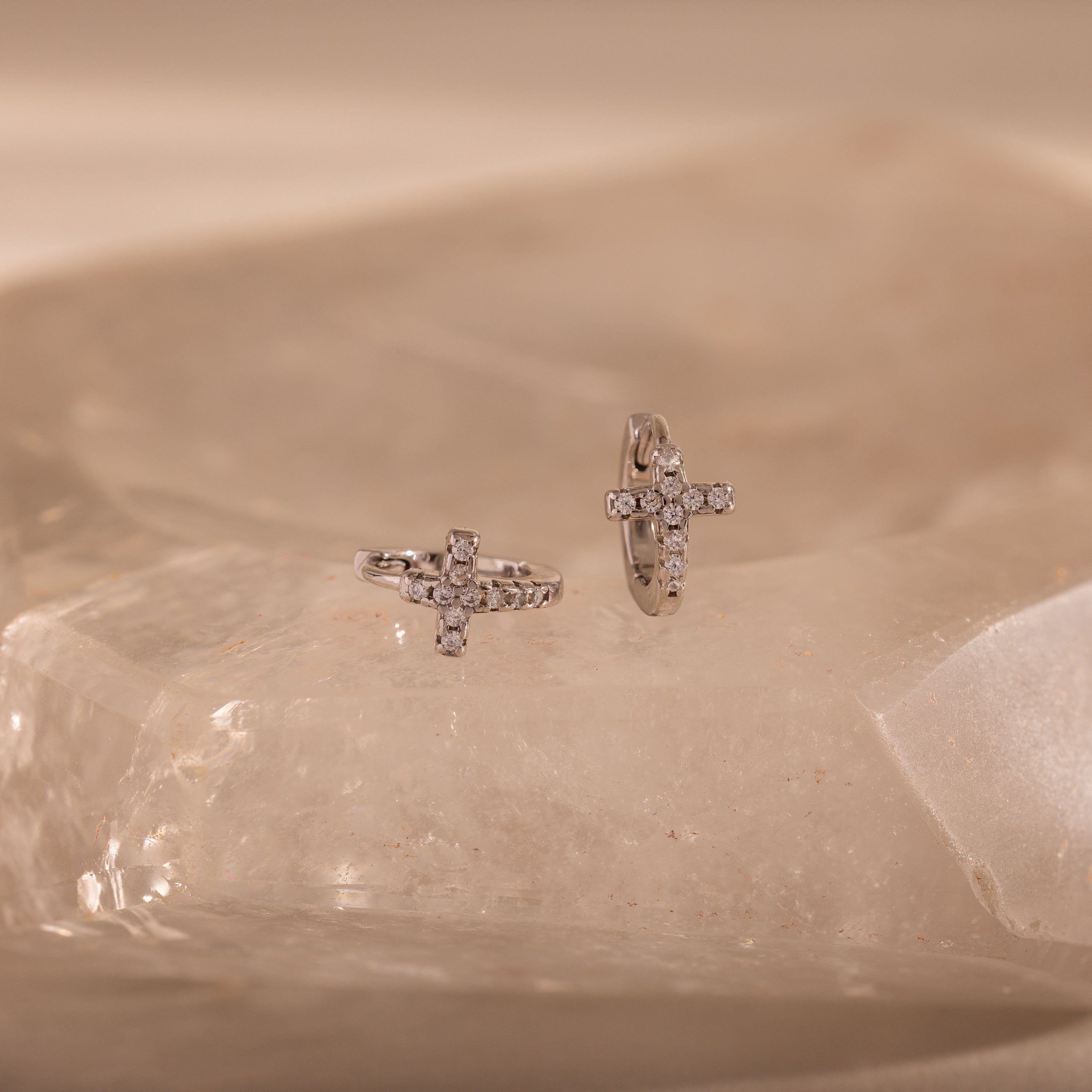 Diamond Cross Huggies in Silver with pavé stones are displayed on a translucent, beige crystal surface.