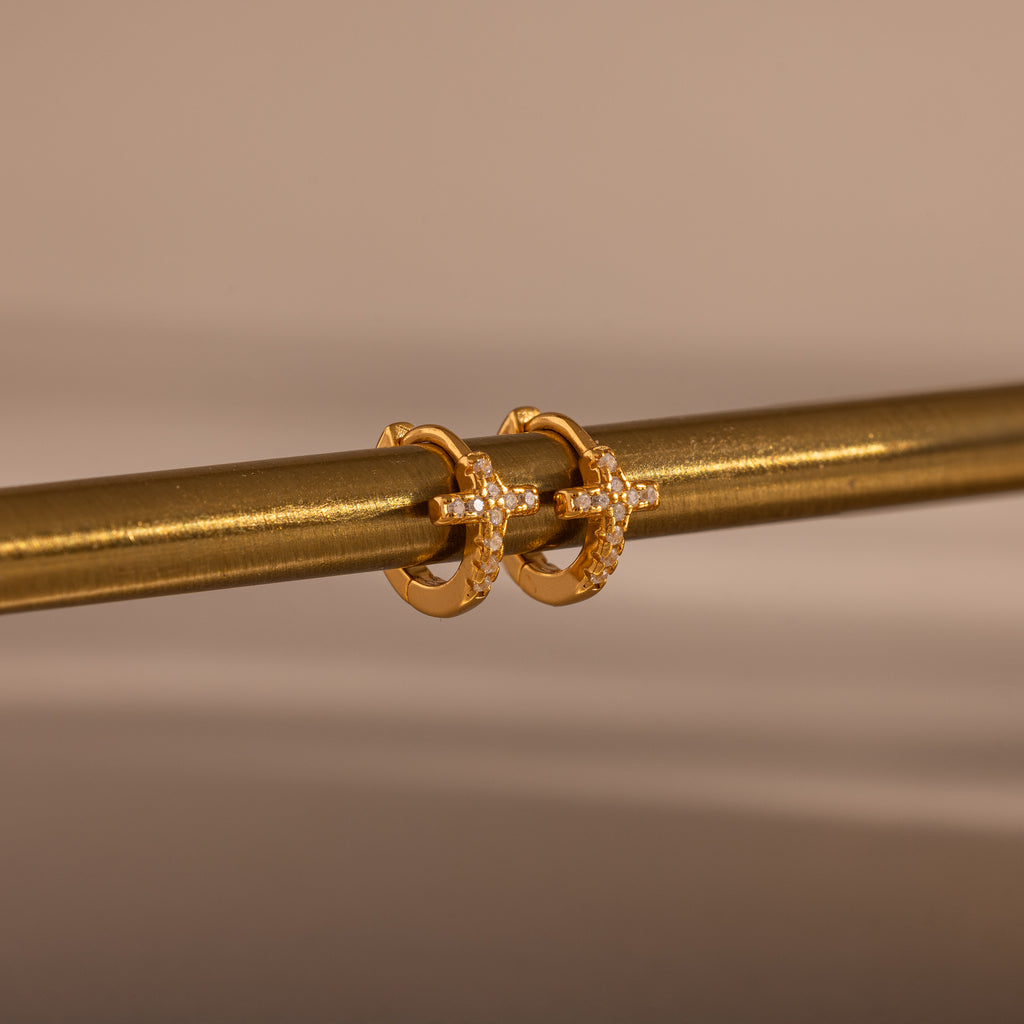 Diamond Cross Huggies in 18K Gold, featuring cross designs and pavé stones, hang on a metallic rod against a neutral background.