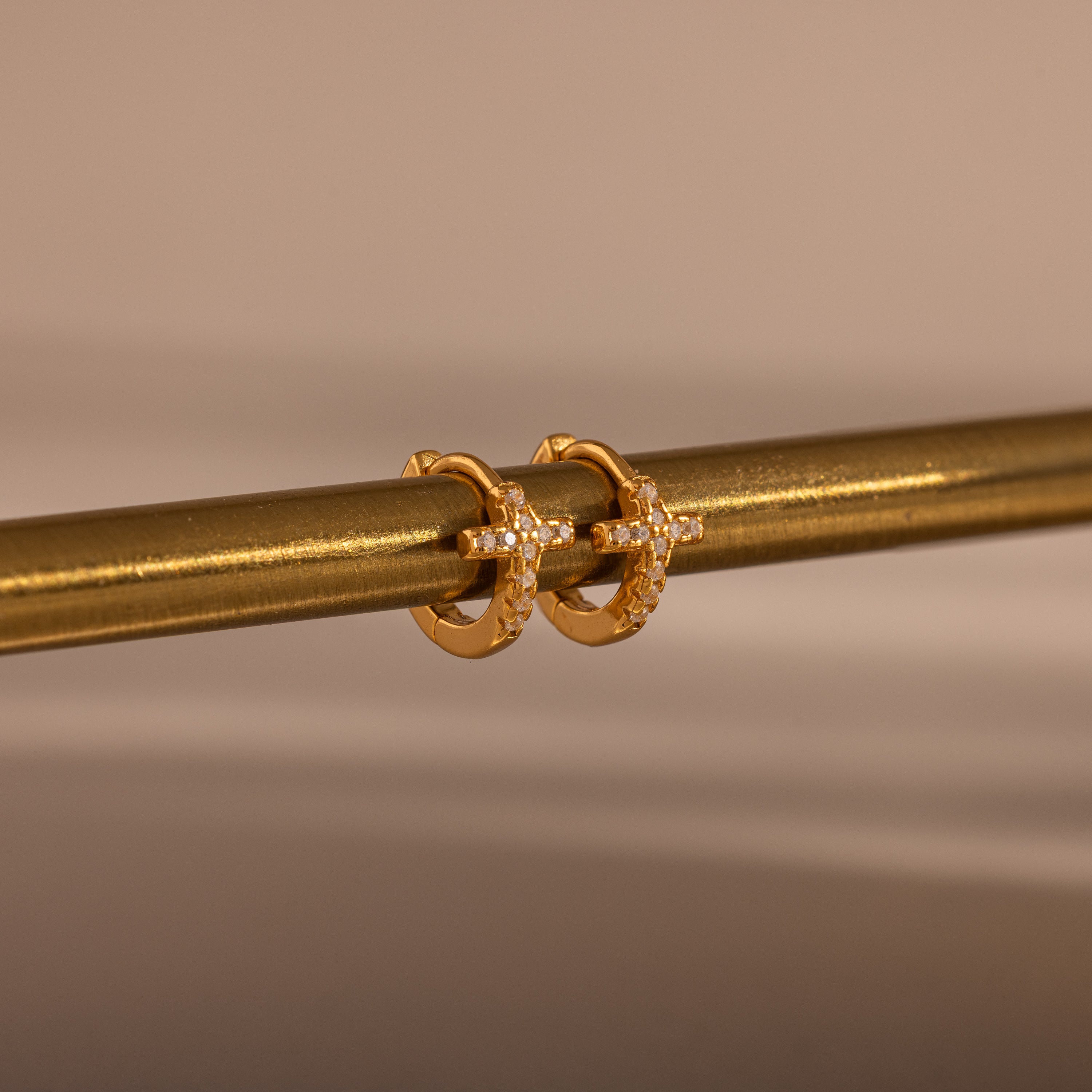 Diamond Cross Huggies in 18K Gold, featuring cross designs and pavé stones, hang on a metallic rod against a neutral background.