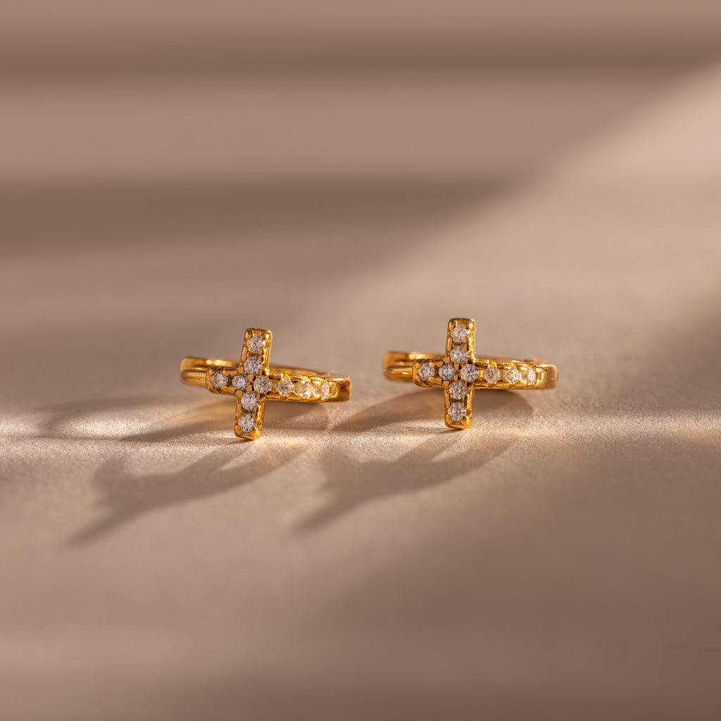 Diamond Cross Huggies in 18K Gold, featuring cross-shaped, diamond-studded designs and pavé stones, rest on a neutral surface with soft shadows.
