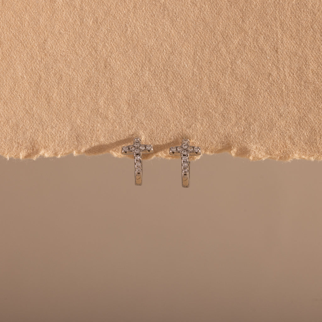 Diamond Cross Huggies in Silver sparkle with pavé stones, displayed against a textured beige background.