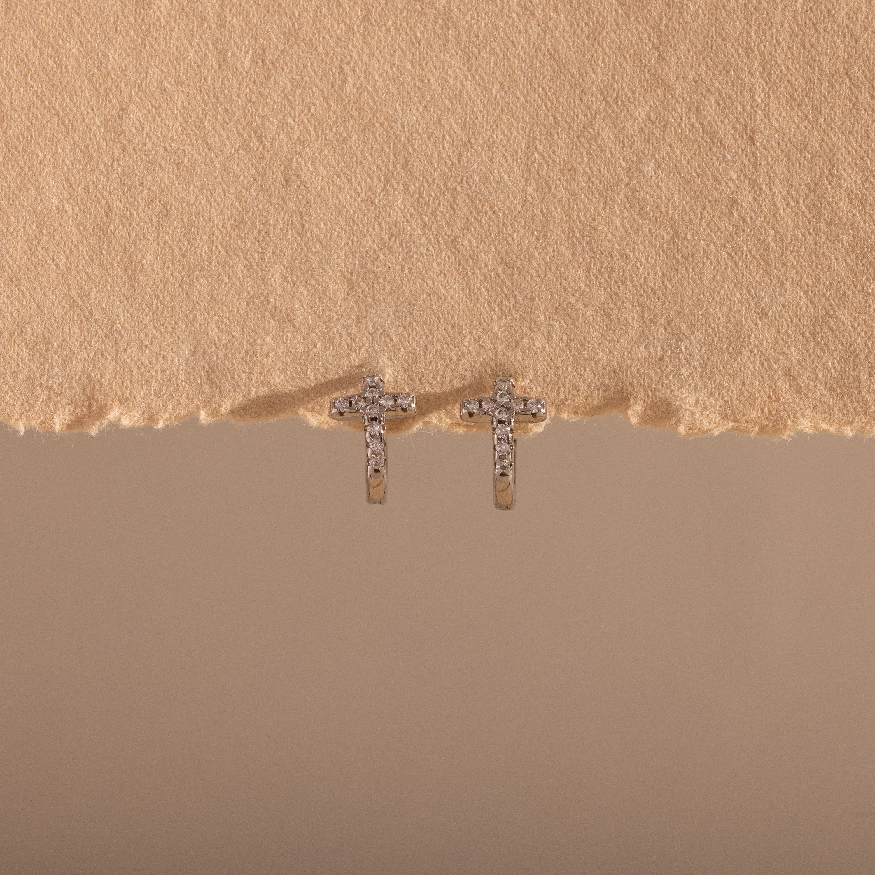 The Diamond Cross Huggies, featuring small silver crosses with pavé stones, are displayed against a textured beige background.