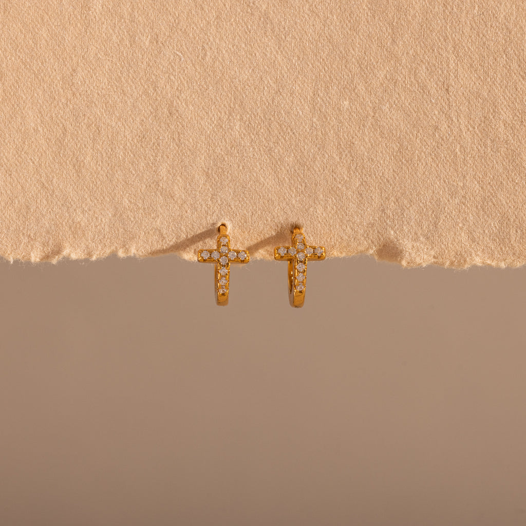 The Diamond Cross Huggies in 18K Gold feature pavé stones forming cross charms—your signature pair, shown against a textured beige background.