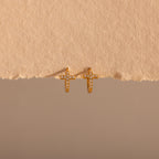 The Diamond Cross Huggies in 18K Gold feature pavé stones forming cross charms—your signature pair, shown against a textured beige background.