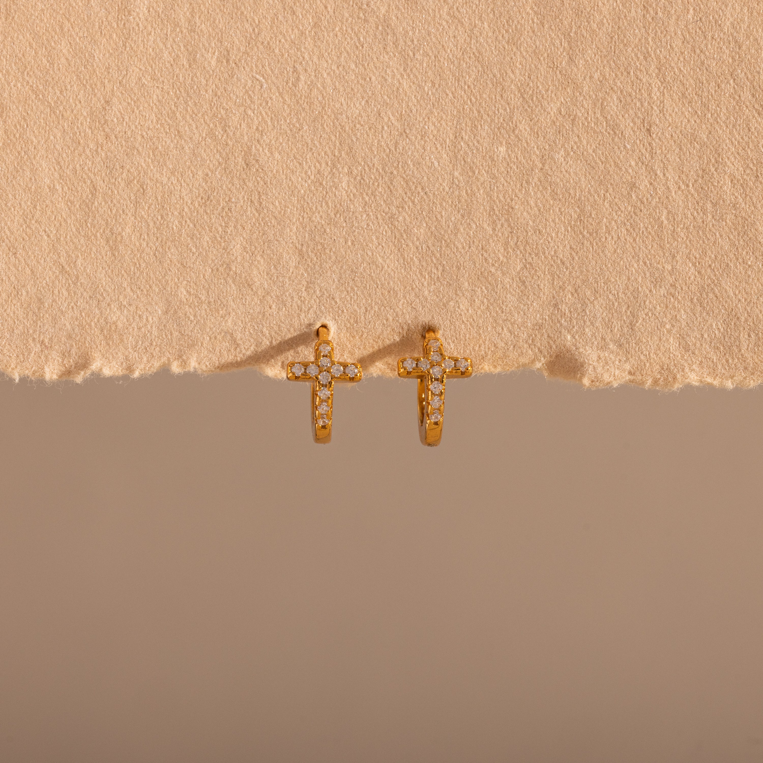The Diamond Cross Huggies in 18K Gold feature pavé stones forming cross charms—your signature pair, shown against a textured beige background.