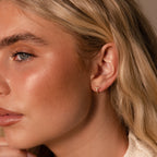 Close-up of a woman with blonde hair wearing the Diamond Cross Huggies in 18K Gold, sparkling subtly in her left ear.
