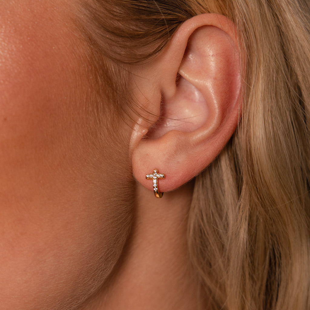 A close-up shows an ear wearing the Diamond Cross Huggies—a small gold hoop with pavé stones on a delicate cross charm, perfect for lovers of dainty, sparkling jewelry.