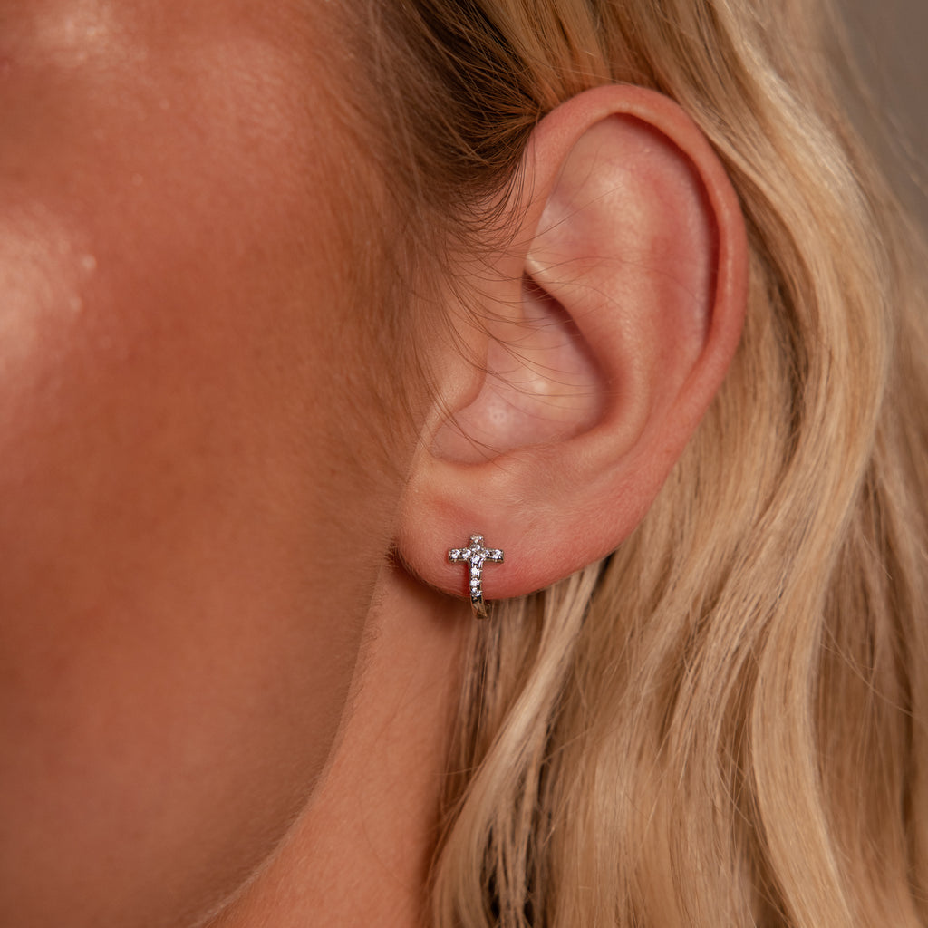 Close-up of a woman's ear wearing Diamond Cross Huggies in Silver adorned with pavé stones, with her blonde hair visible.