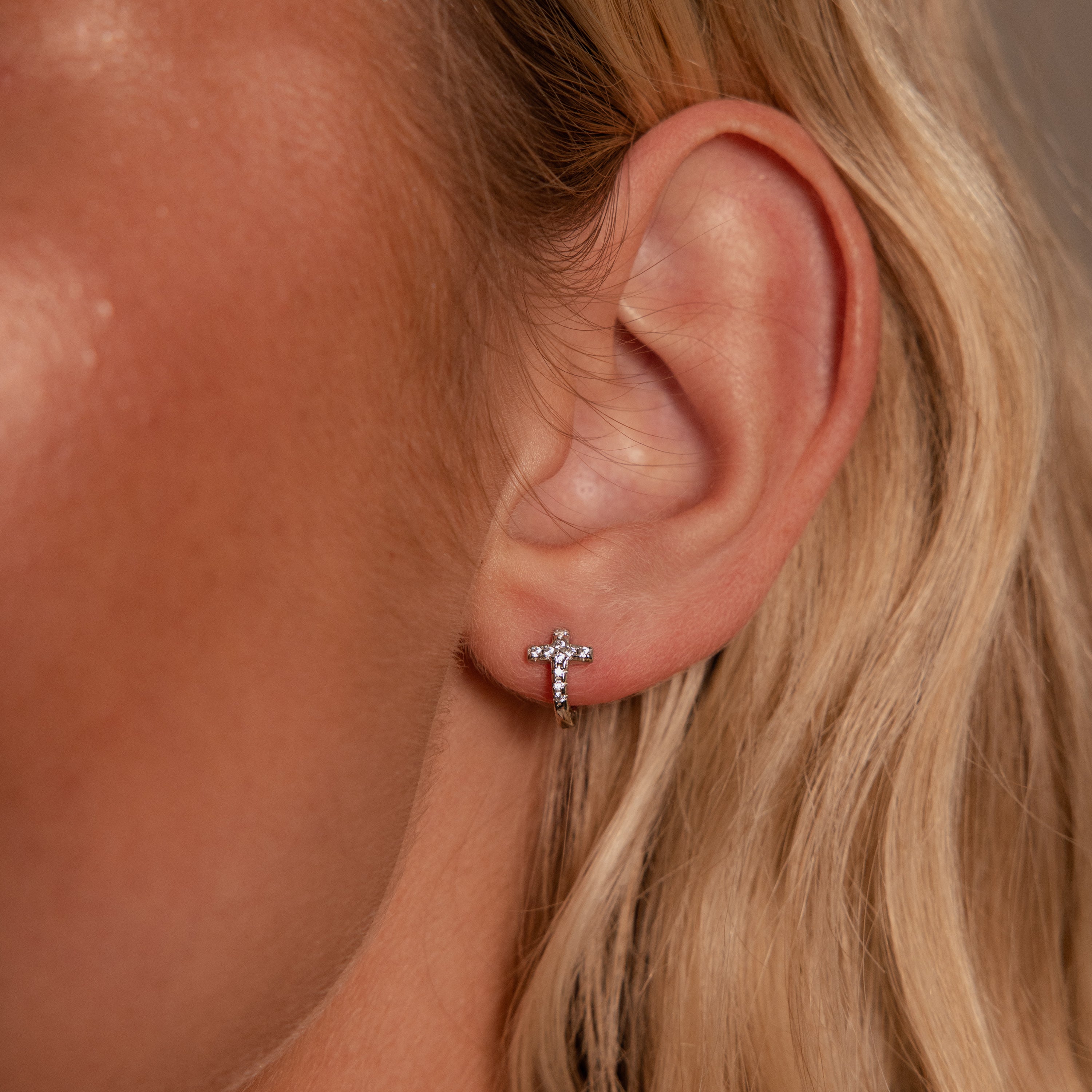 Close-up of a woman's ear wearing Diamond Cross Huggies in Silver adorned with pavé stones, with her blonde hair visible.