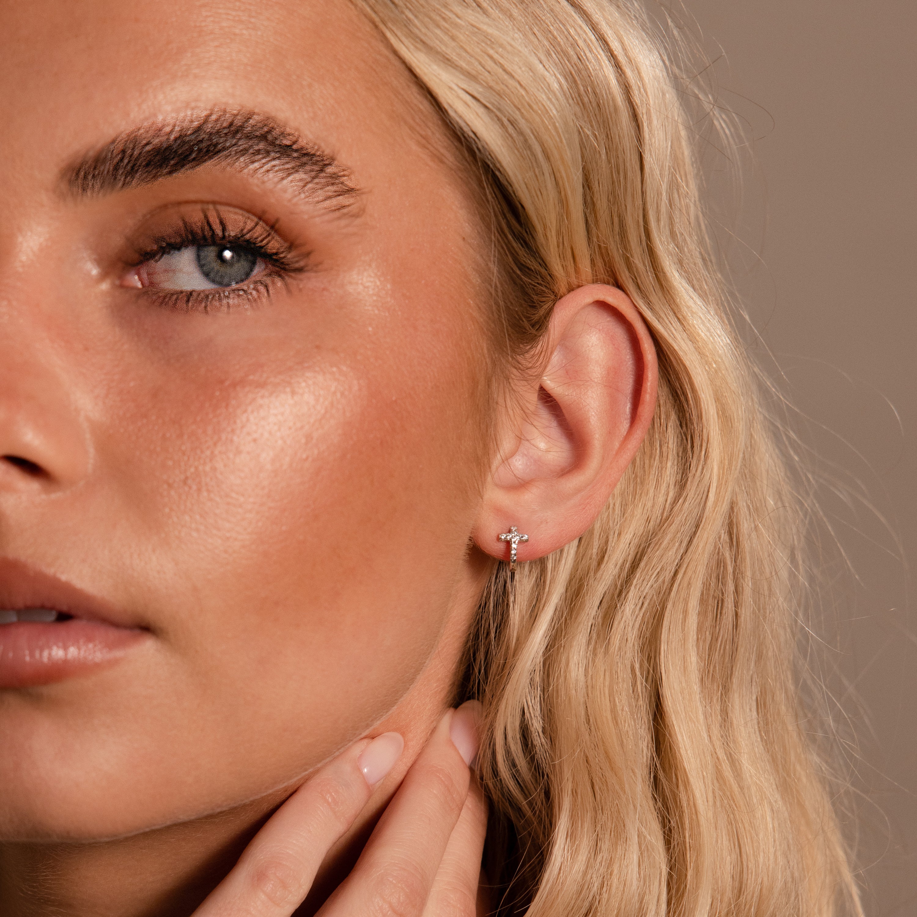 A woman with blonde hair wears Diamond Cross Huggies, featuring pavé stones, as she touches her neck against a neutral background.