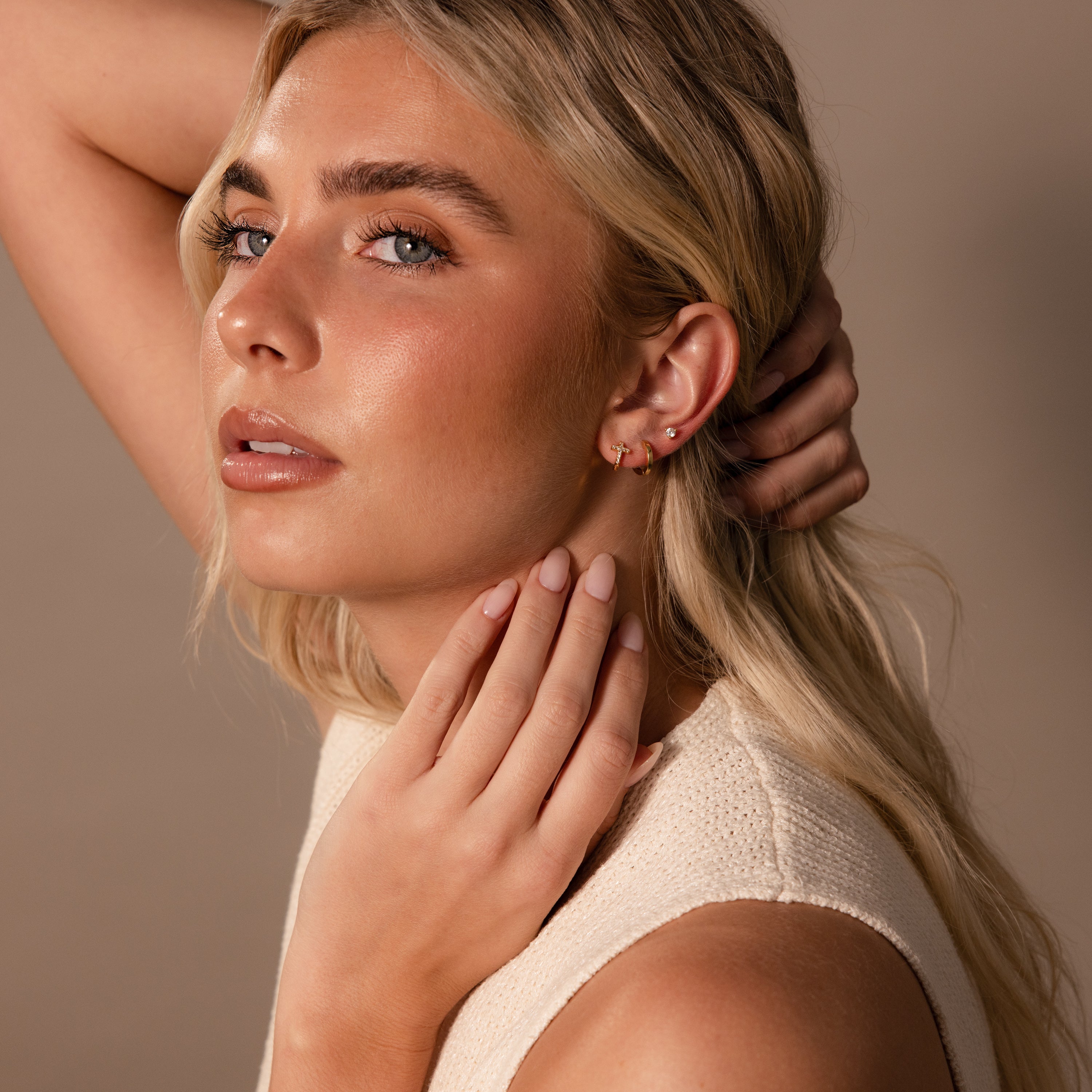 A blonde woman with neutral makeup and a cream sleeveless top poses with her hand on her neck, wearing Diamond Cross Huggies in 18K Gold adorned with pavé stones.