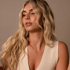 Woman with long wavy blonde hair wearing a cream knit top and layered gold necklaces, including a minimalist necklace and the Vintage Cross Necklace in 18K Gold, gazes to the side.