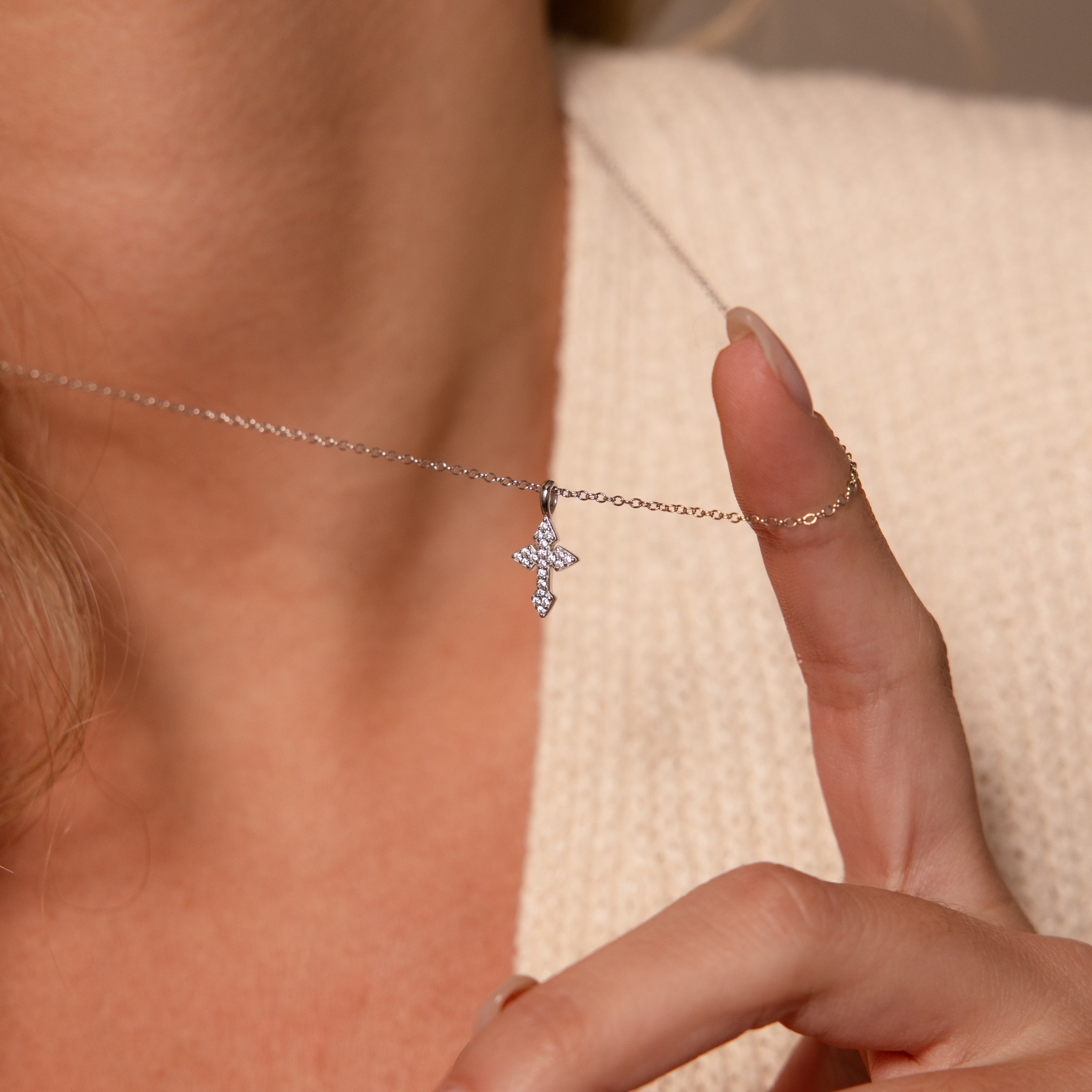 Someone wearing a cream sweater holds the Vintage Cross Necklace in Silver, featuring a small, sparkling pavé-finish pendant, against their neck.
