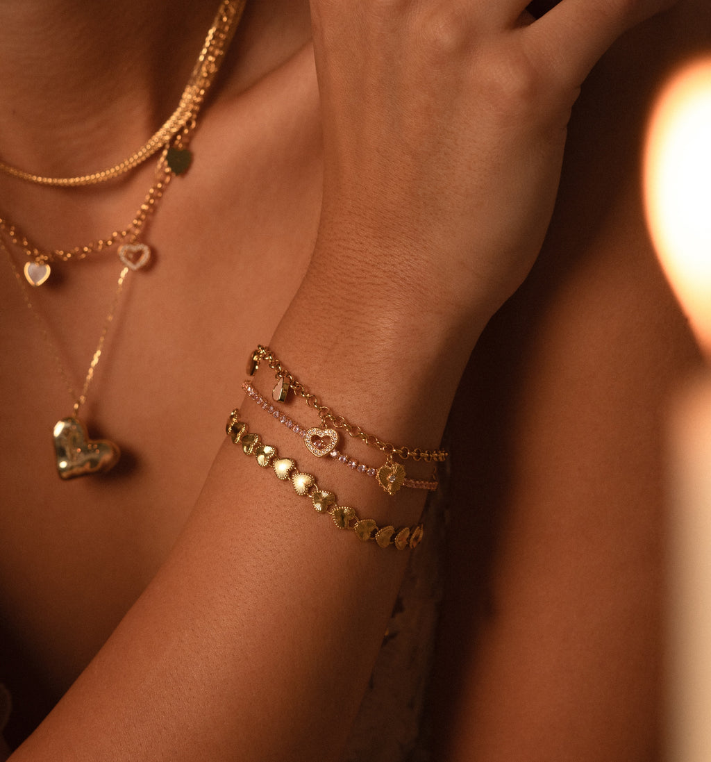 A woman wearing layered gold necklaces and a Heart Charm Bracelet with disc charms touches her shoulder—the perfect gift for your girlfriend.