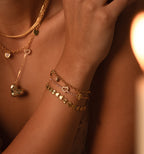 A woman wearing layered gold necklaces and a Heart Charm Bracelet with disc charms touches her shoulder—the perfect gift for your girlfriend.