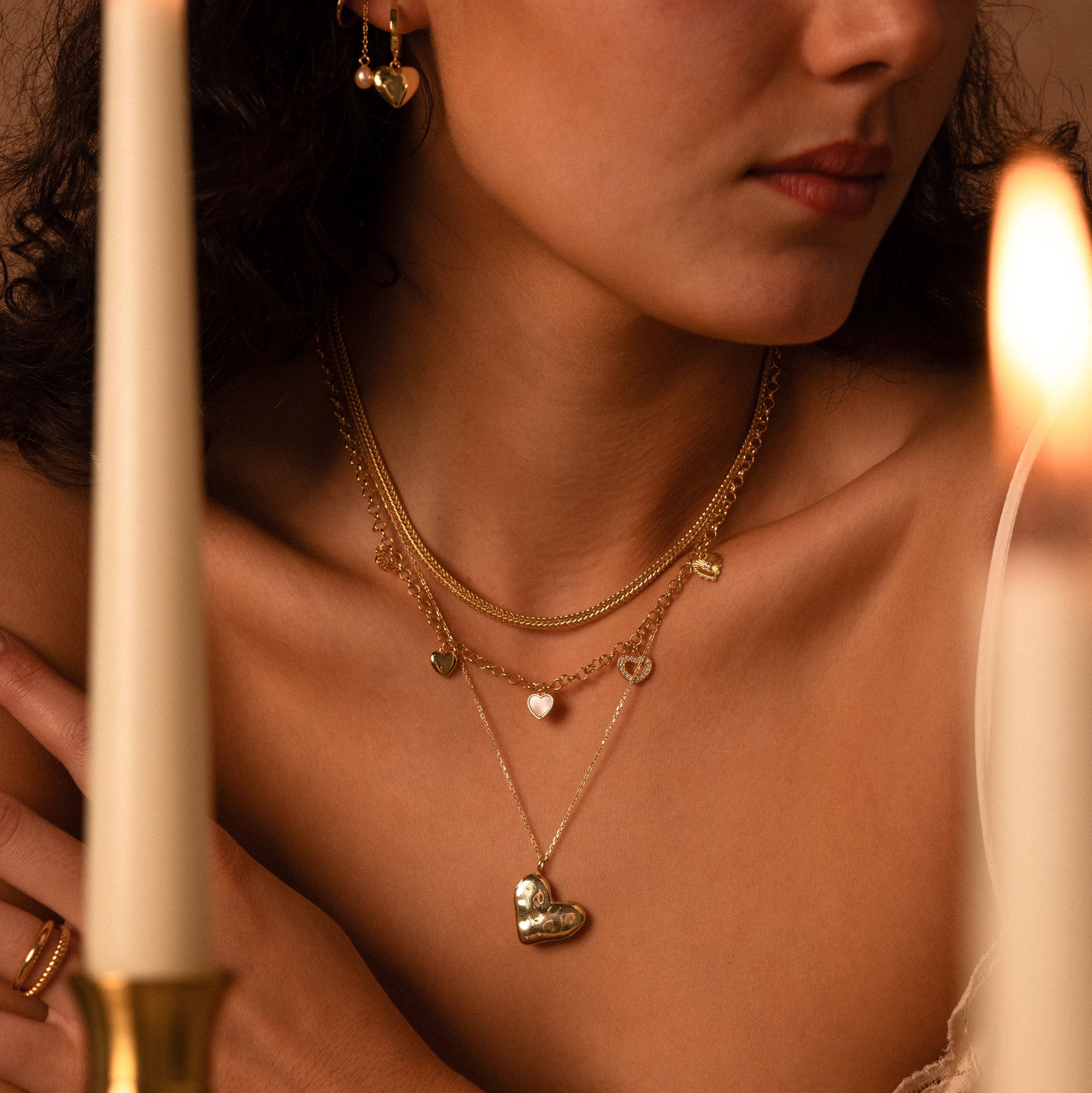 A woman wears the Heart Charm Necklace in 18K Gold, layered with delicate heart accents and matching earrings, with candles glowing in the foreground.