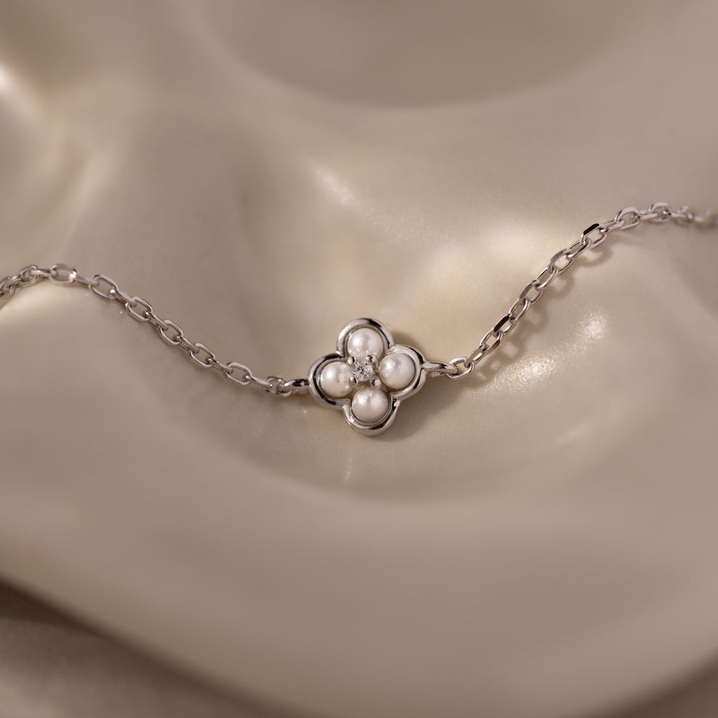 The Rounded Pearl Flower Bracelet in Sterling Silver features a four-petal floral charm, each petal adorned with a small pearl, creating an elegant accessory showcased on a beige surface.