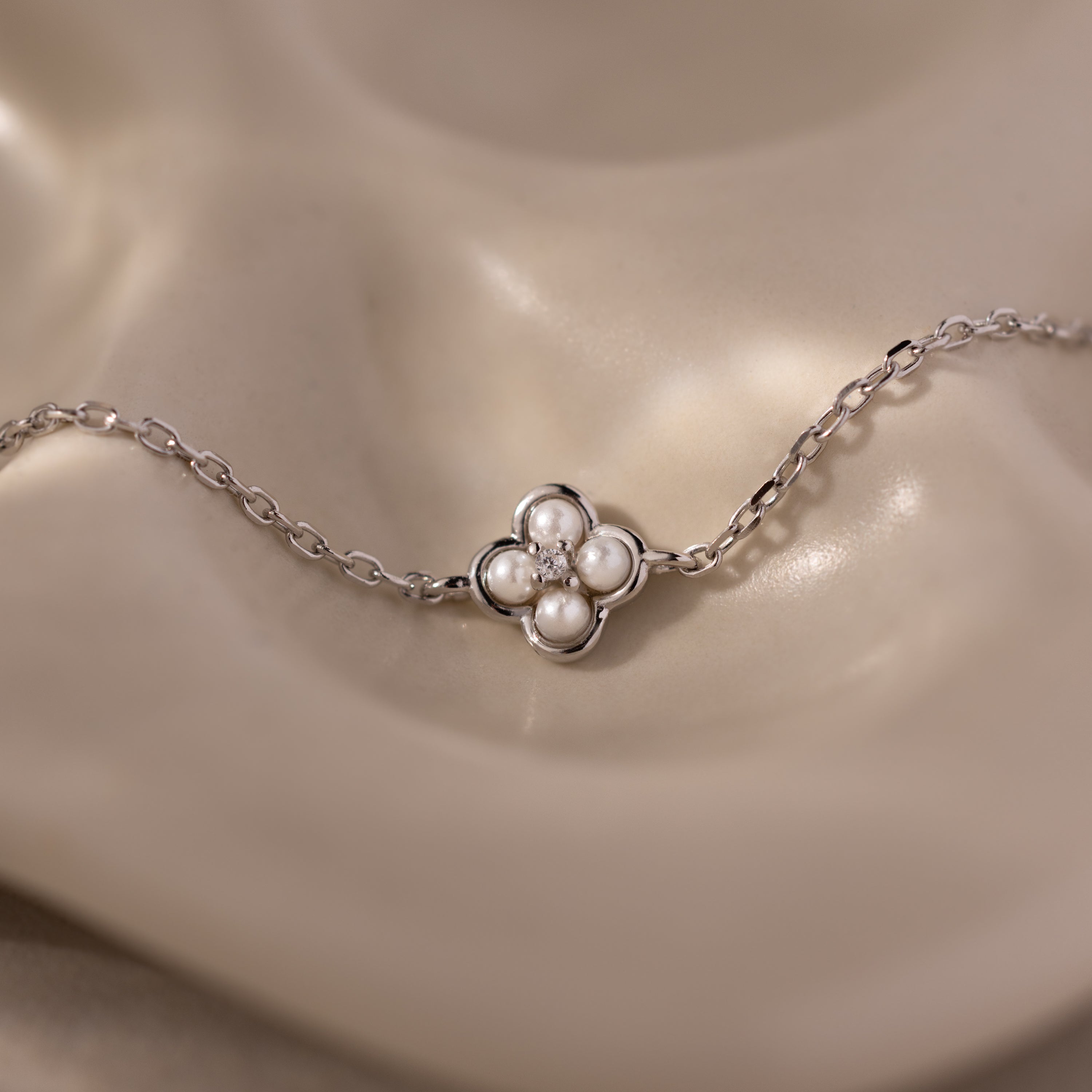 The Rounded Pearl Flower Bracelet in Sterling Silver features a four-petal floral charm, each petal adorned with a small pearl, creating an elegant accessory showcased on a beige surface.