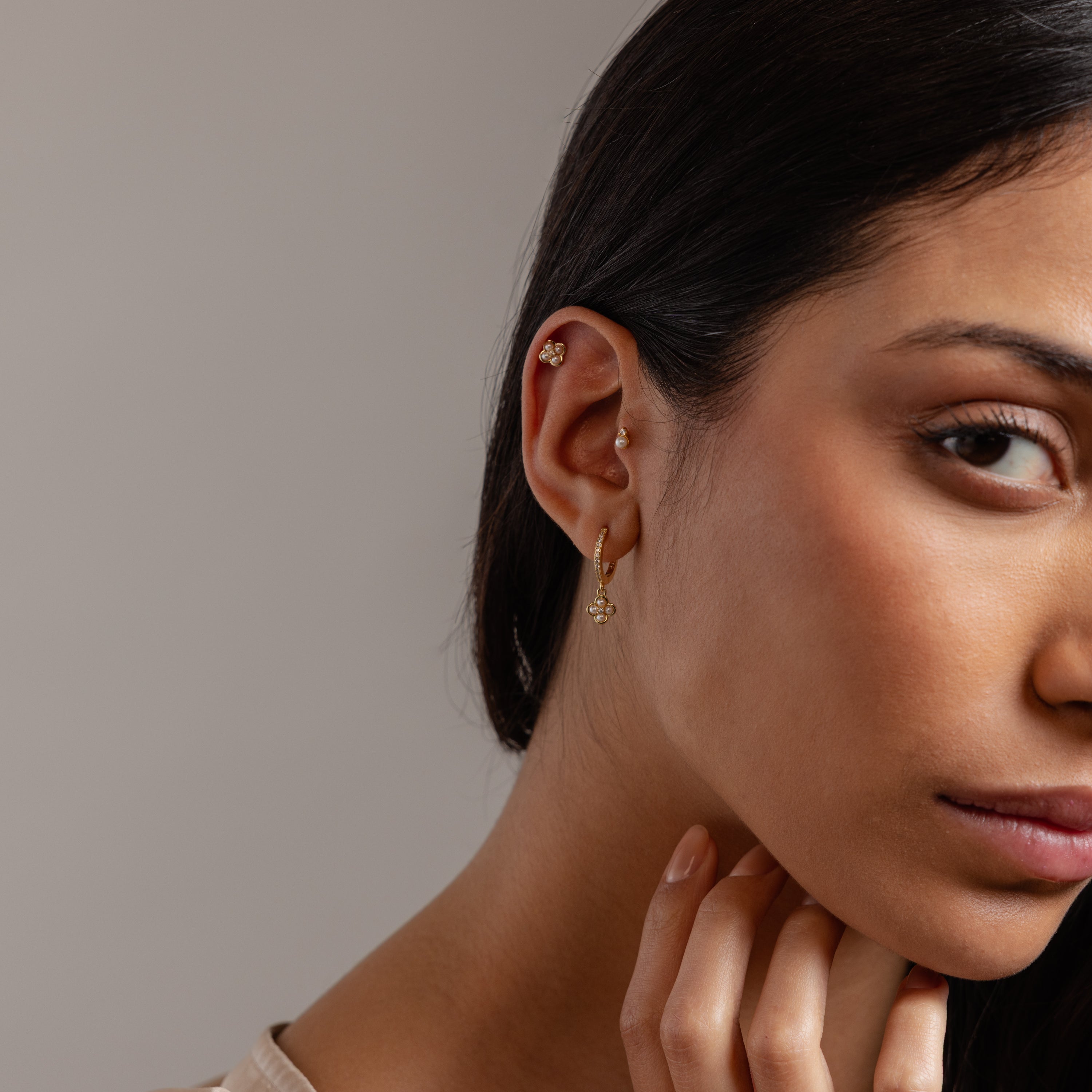 Close-up of a woman wearing multiple earrings, featuring the Pave Rounded Pearl Flower Hoops in 18K Gold, with her hand touching her chin.