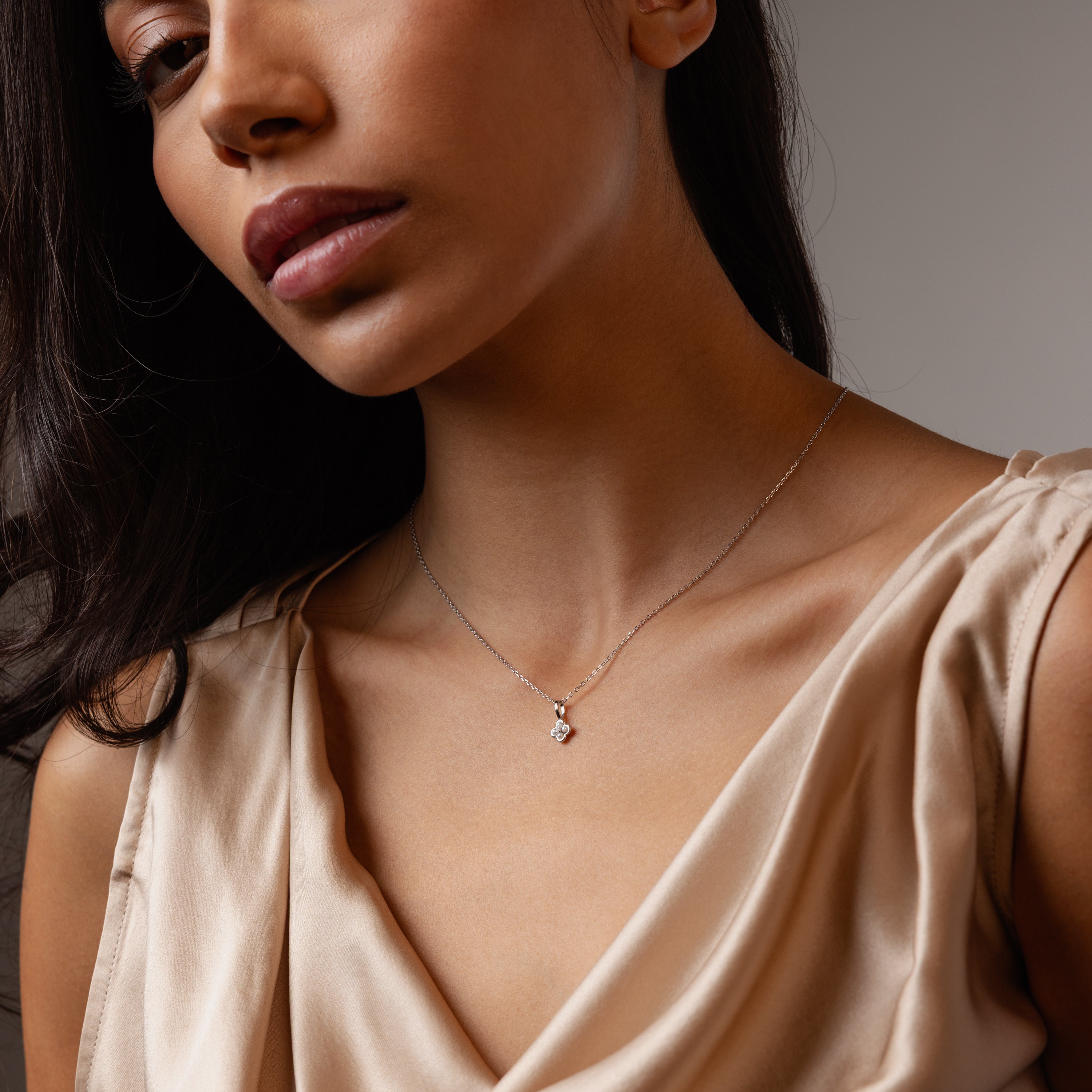 A woman in a beige satin top wears the Rounded Pearl Flower Necklace in Sterling Silver, its small pendant shining against a neutral background—perfect for everyday layering.