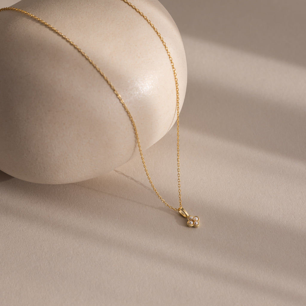 The Rounded Pearl Flower Necklace in 18K Gold features a delicate floral pendant, elegantly displayed on a smooth, beige stone—ideal for lovers of refined floral jewelry.
