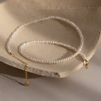 The Summer Pearl Beaded Necklace in 18K Gold features delicate pearl beads and a gold clasp, shown on a textured beige background—a stylish modern pearl jewelry piece ideal for summer.