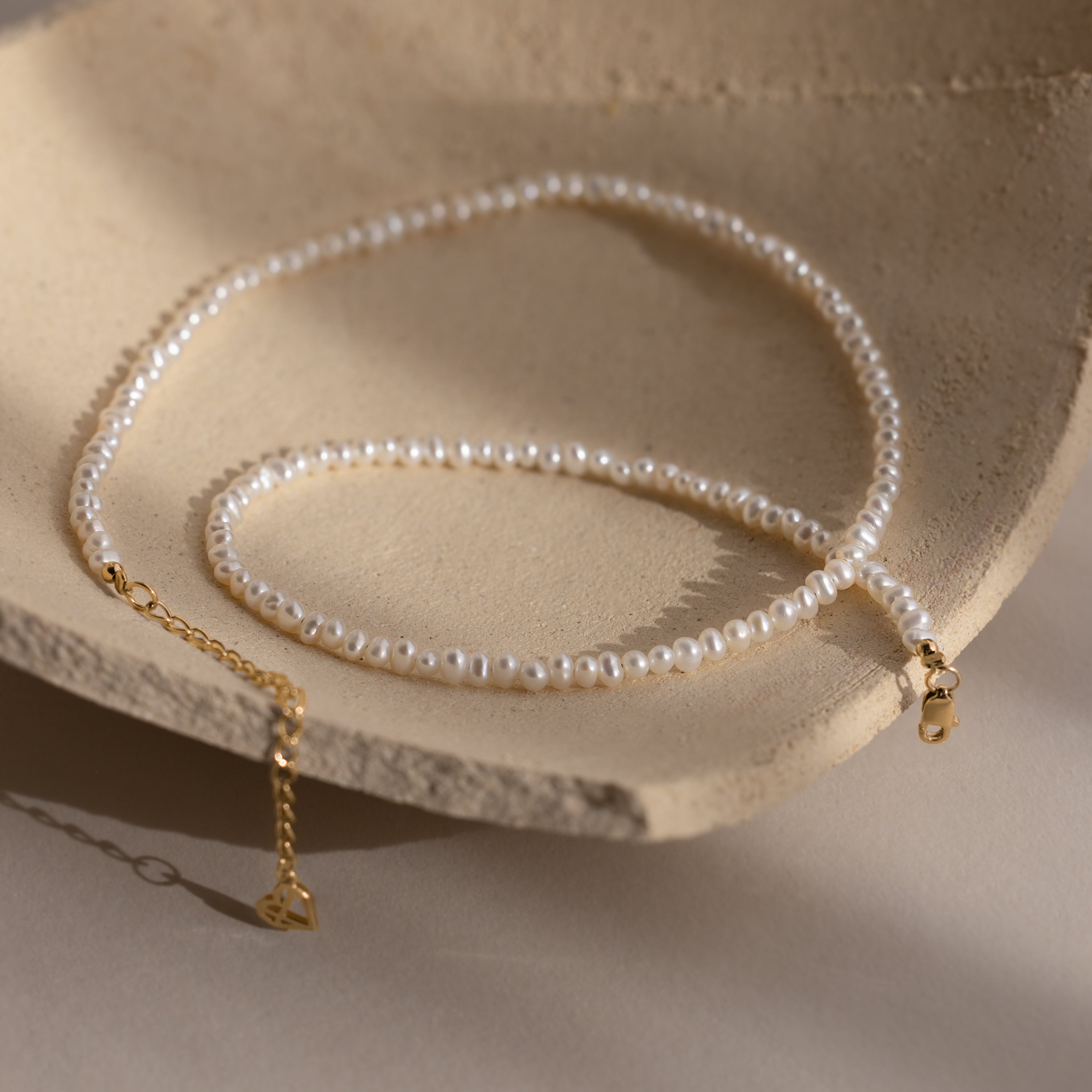 The Summer Pearl Beaded Necklace in 18K Gold features delicate pearl beads and a gold clasp, shown on a textured beige background—a stylish modern pearl jewelry piece ideal for summer.