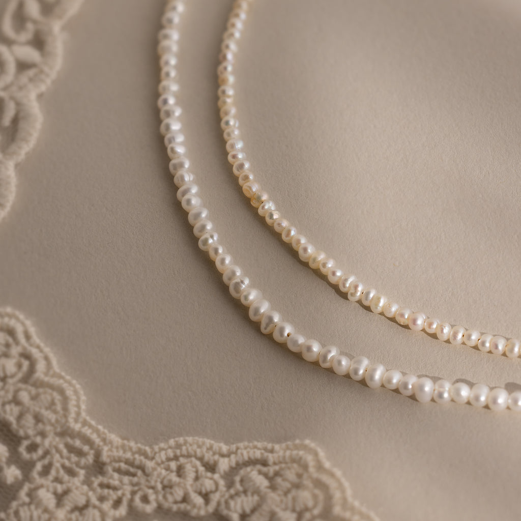 The Summer Pearl Beaded Necklace in 18K Gold rests on a beige surface with lace corners—a modern pearl jewelry piece, perfect for summer.