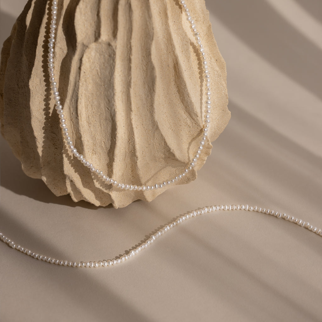 The Summer Pearl Beaded Necklace in 18K Gold drapes elegantly over a textured beige sculpture, casting gentle shadows—a chic and modern pearl jewelry piece.