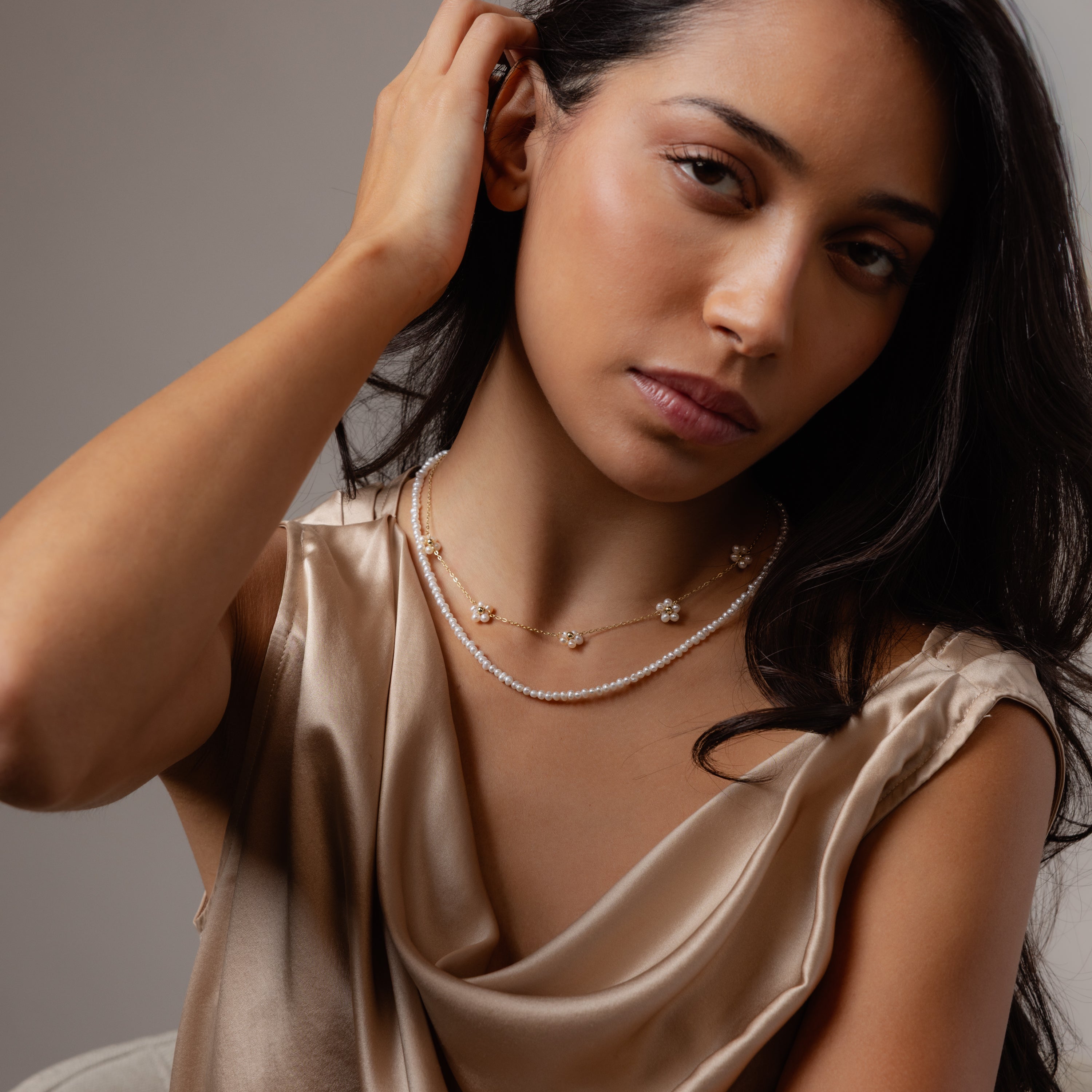 A woman in a silky beige top wears the Summer Pearl Beaded Necklace in 18K Gold, styled with layered pearls, touching her hair and looking at the camera.