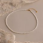 The Summer Pearl Beaded Necklace in 18K Gold features delicate pearls with a gold clasp, displayed on beige fabric beside lace—a chic modern jewelry piece for summer.