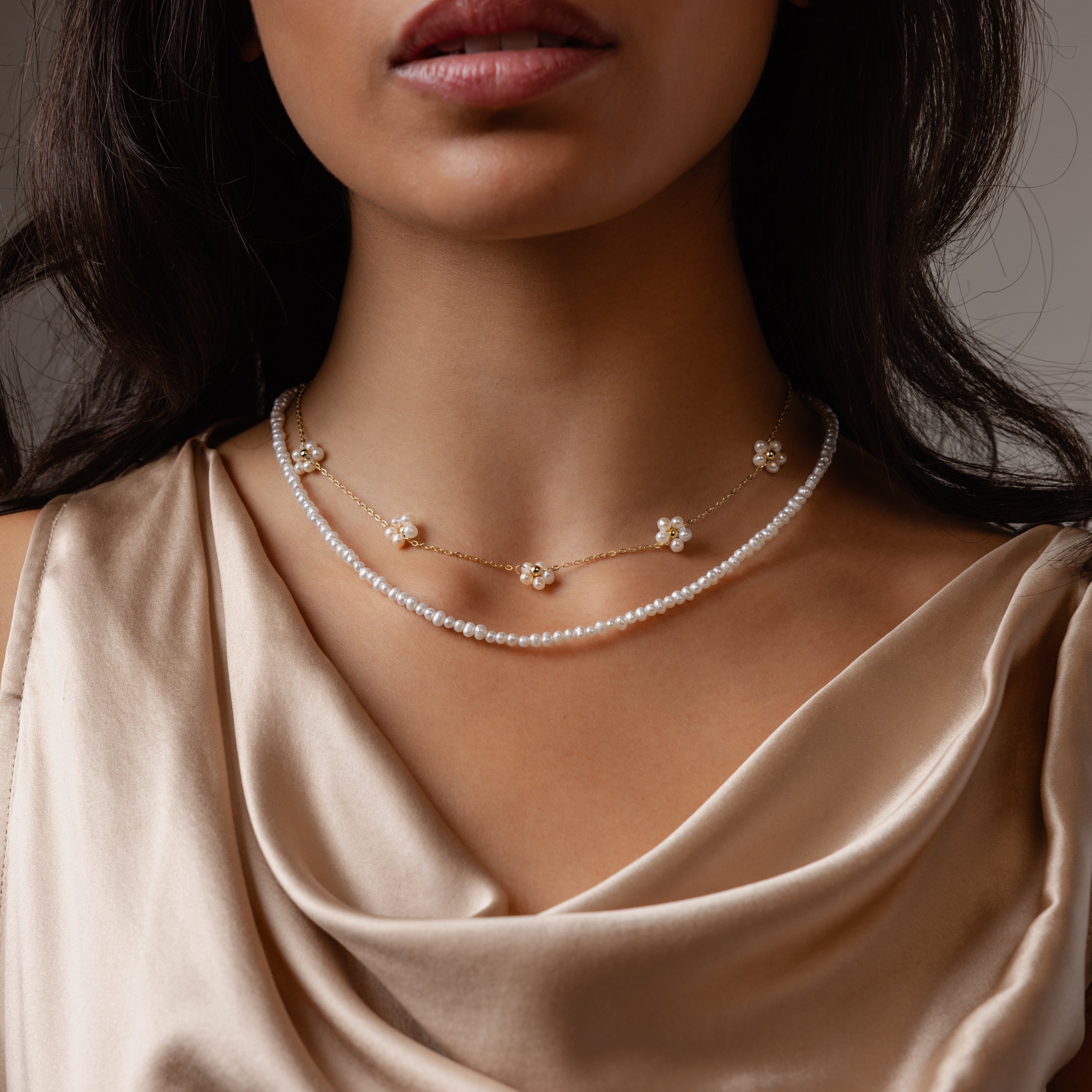 A woman wears the Summer Pearl Beaded Necklace in 18K Gold, layered with gold floral pieces and a champagne satin top—perfect summer jewelry.