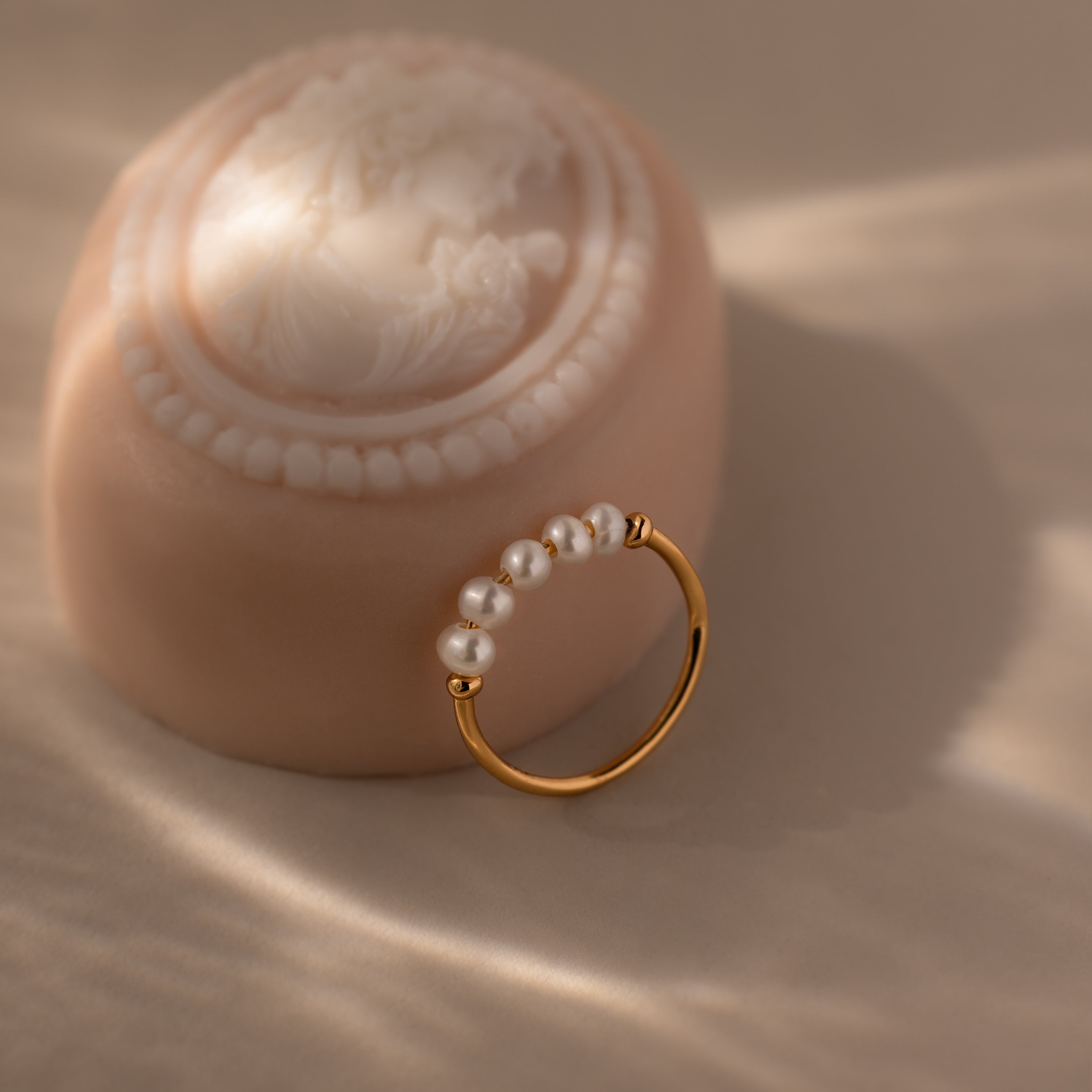 The Beaded Pearl Ring, featuring delicate small pearls, is displayed next to a decorative cameo on a soft beige surface—an elegant showcase of refined jewelry.