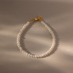 The Venus Pearl Bracelet features freshwater pearls and a gold clasp, displayed in a loose oval on a beige surface with soft lighting—an elegant accessory perfect for any occasion.