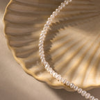 The Venus Pearl Bracelet, crafted from freshwater pearls, rests on a ceramic shell-shaped dish and casts gentle shadows across the surface.