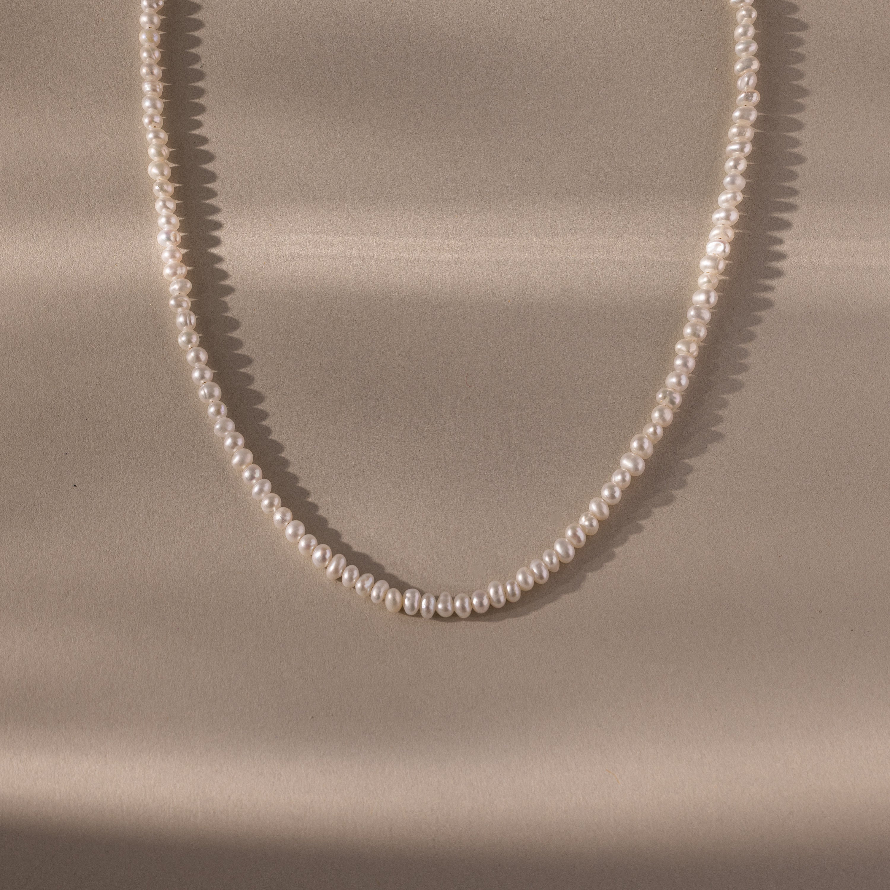 The Venus Pearl Necklace features a single strand of small freshwater pearls in a loose oval shape on a beige surface, showcasing the elegance of timeless jewelry.