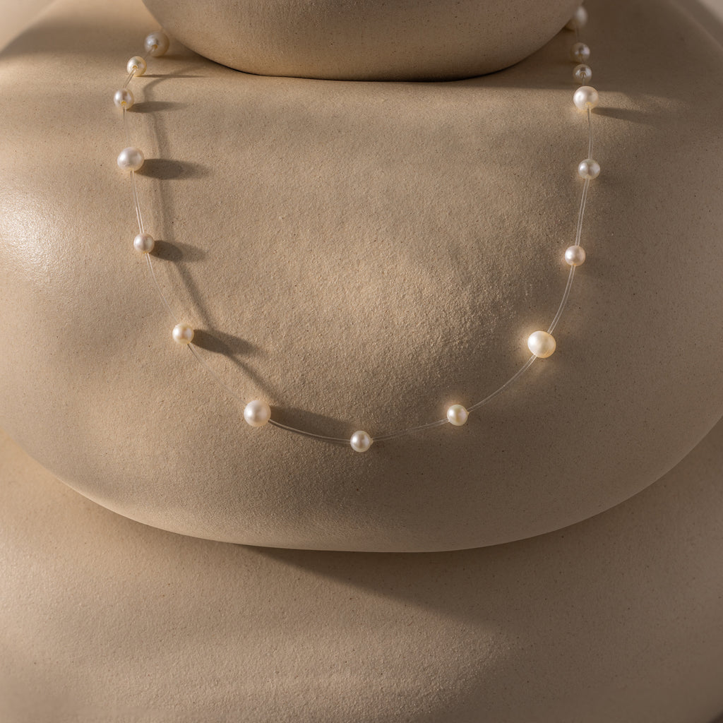 The Floating Pearl Necklace, featuring delicate freshwater pearls, is displayed on smooth beige rounded stones, casting soft shadows.