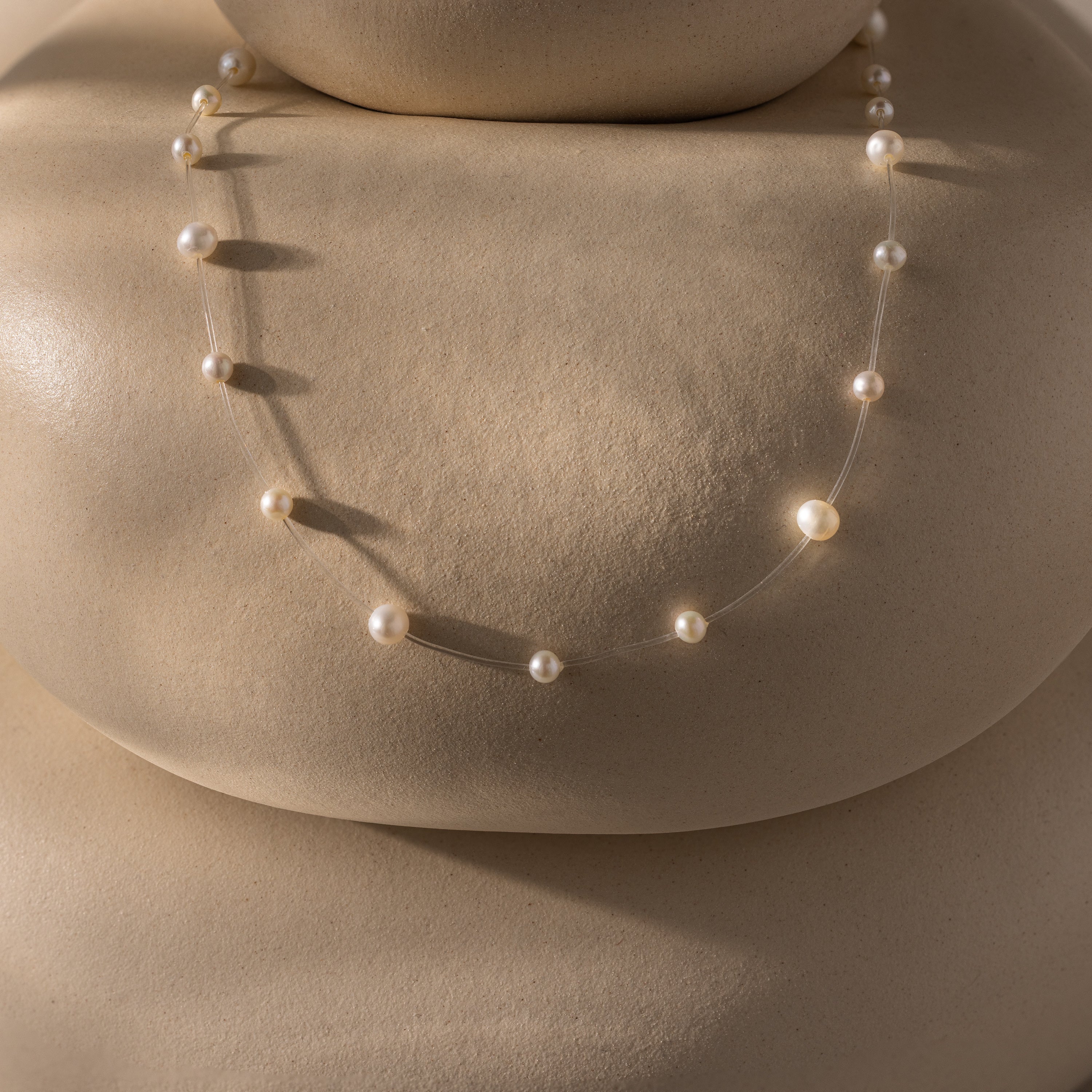 The Floating Pearl Necklace, featuring delicate freshwater pearls, is displayed on smooth beige rounded stones, casting soft shadows.