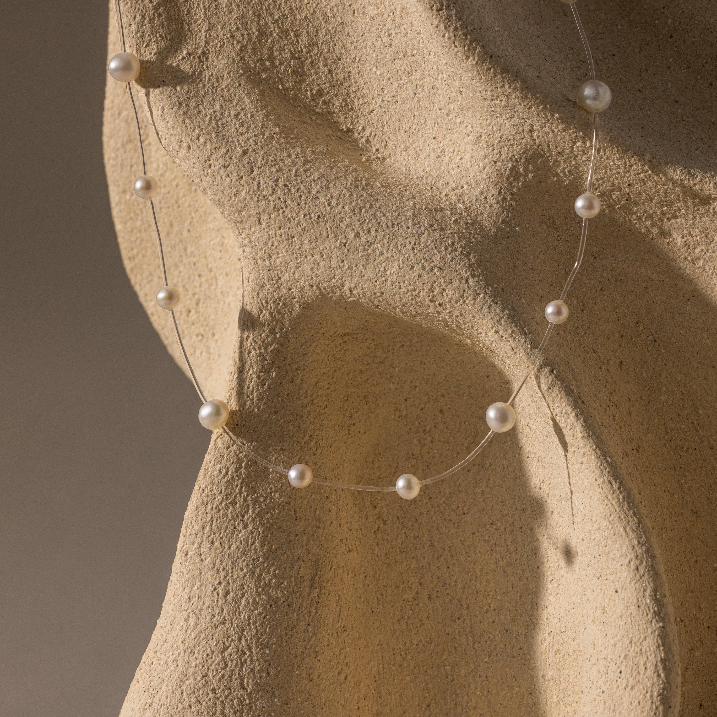 The Floating Pearl Necklace features luminous freshwater pearls gracefully draped over a textured beige surface under soft lighting.
