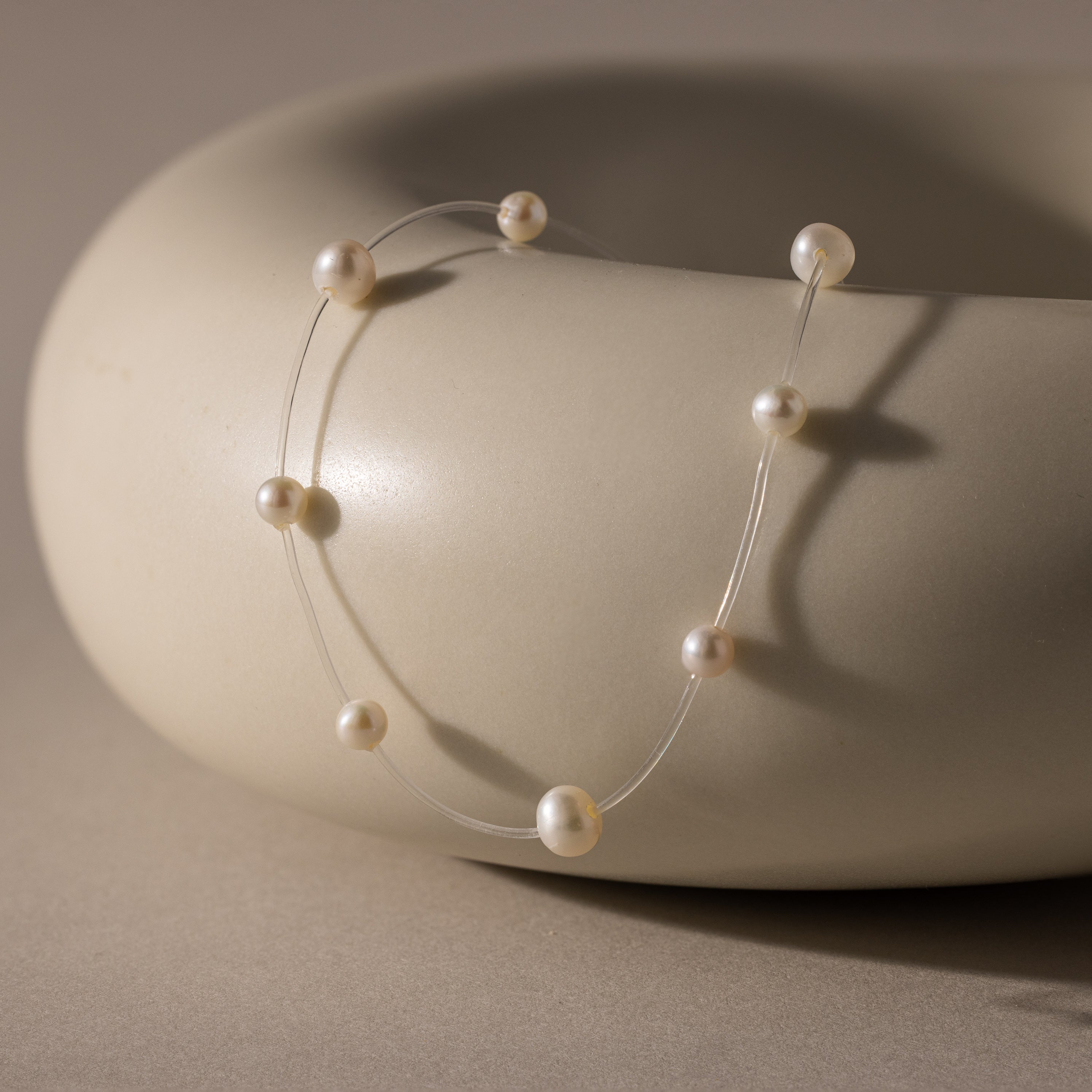 The Floating Pearl Necklace features delicate freshwater pearls on a clear strand, creating a graceful floating effect as it drapes over a smooth, round beige ceramic bowl.