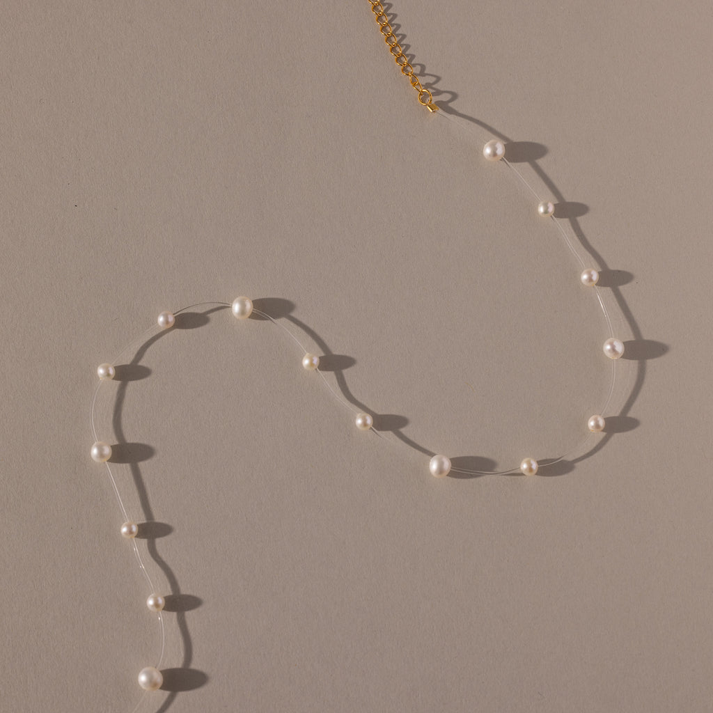 The Floating Pearl Necklace, featuring delicate freshwater pearls and a gold clasp, is arranged in a graceful curve on a beige surface.