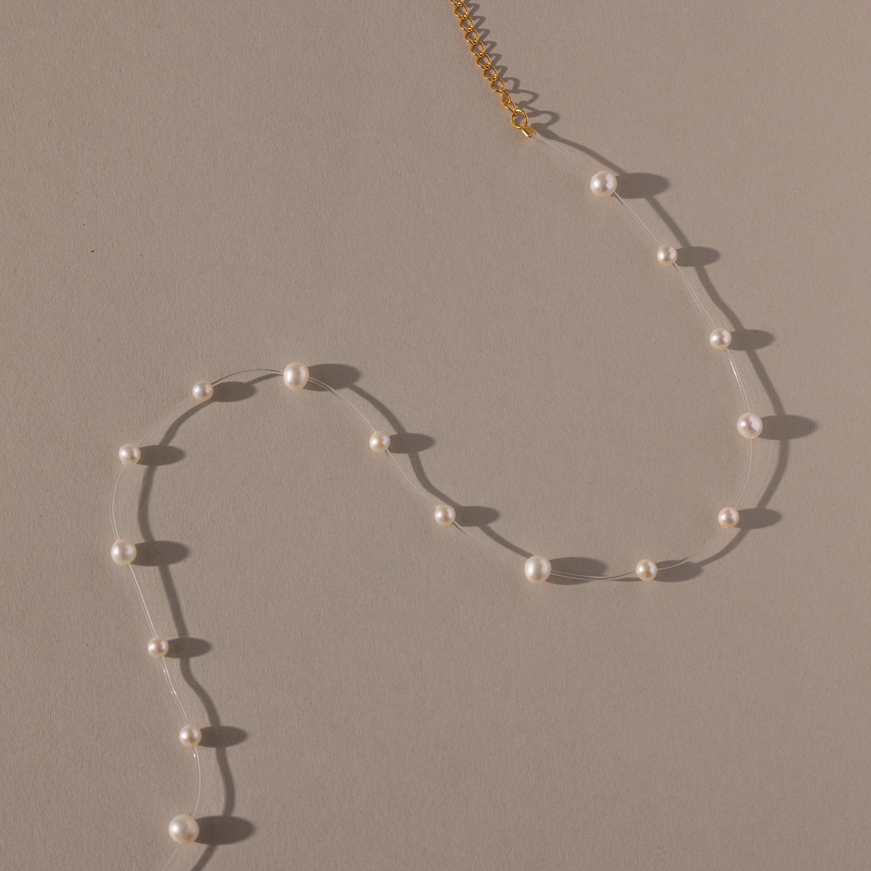 The Floating Pearl Necklace, featuring delicate freshwater pearls and a gold clasp, is arranged in a graceful curve on a beige surface.