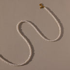 The Venus Pearl Necklace, a single-strand freshwater pearl necklace with a gold clasp, rests on a beige background, showcasing timeless elegance.