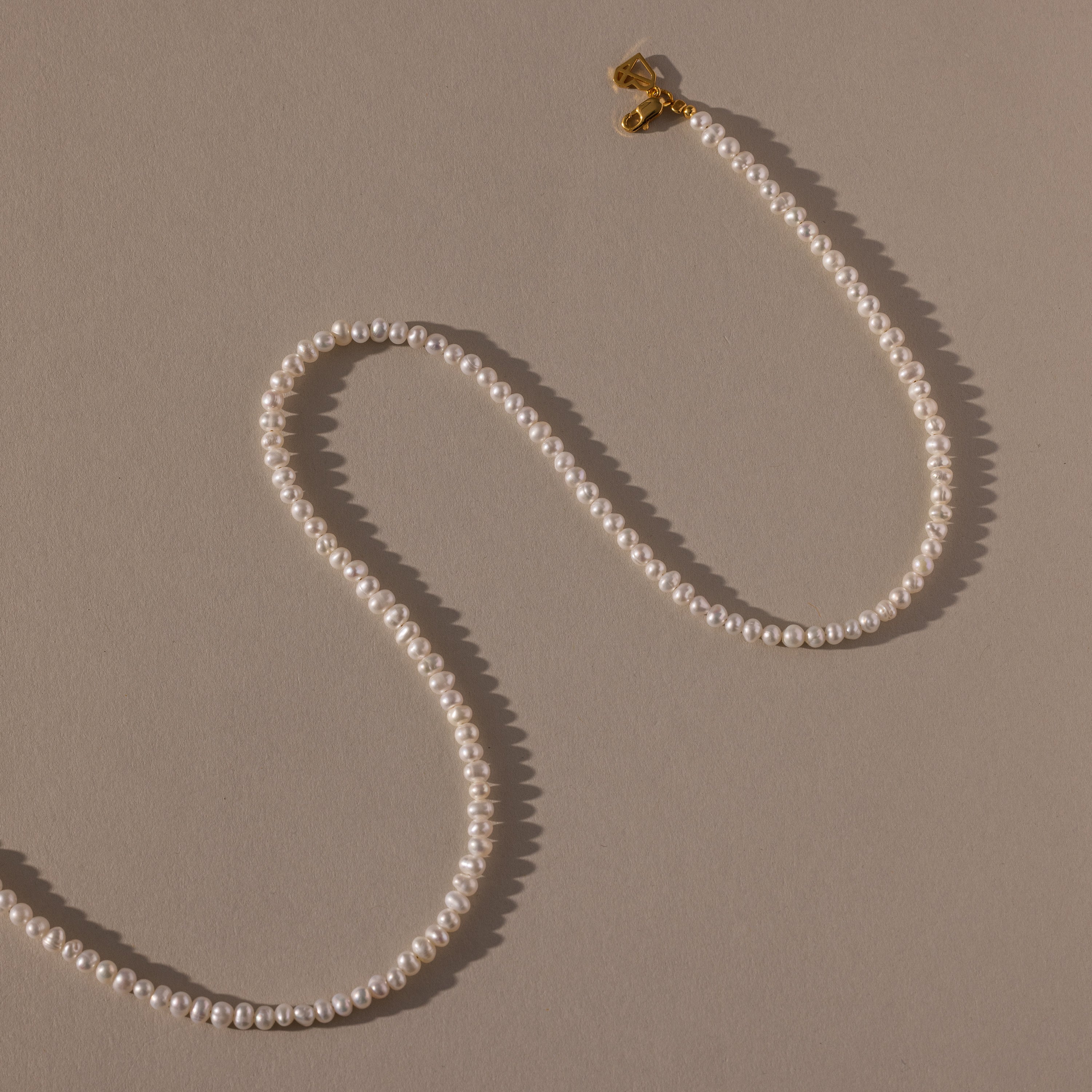 The Venus Pearl Necklace, a single-strand freshwater pearl necklace with a gold clasp, rests on a beige background, showcasing timeless elegance.
