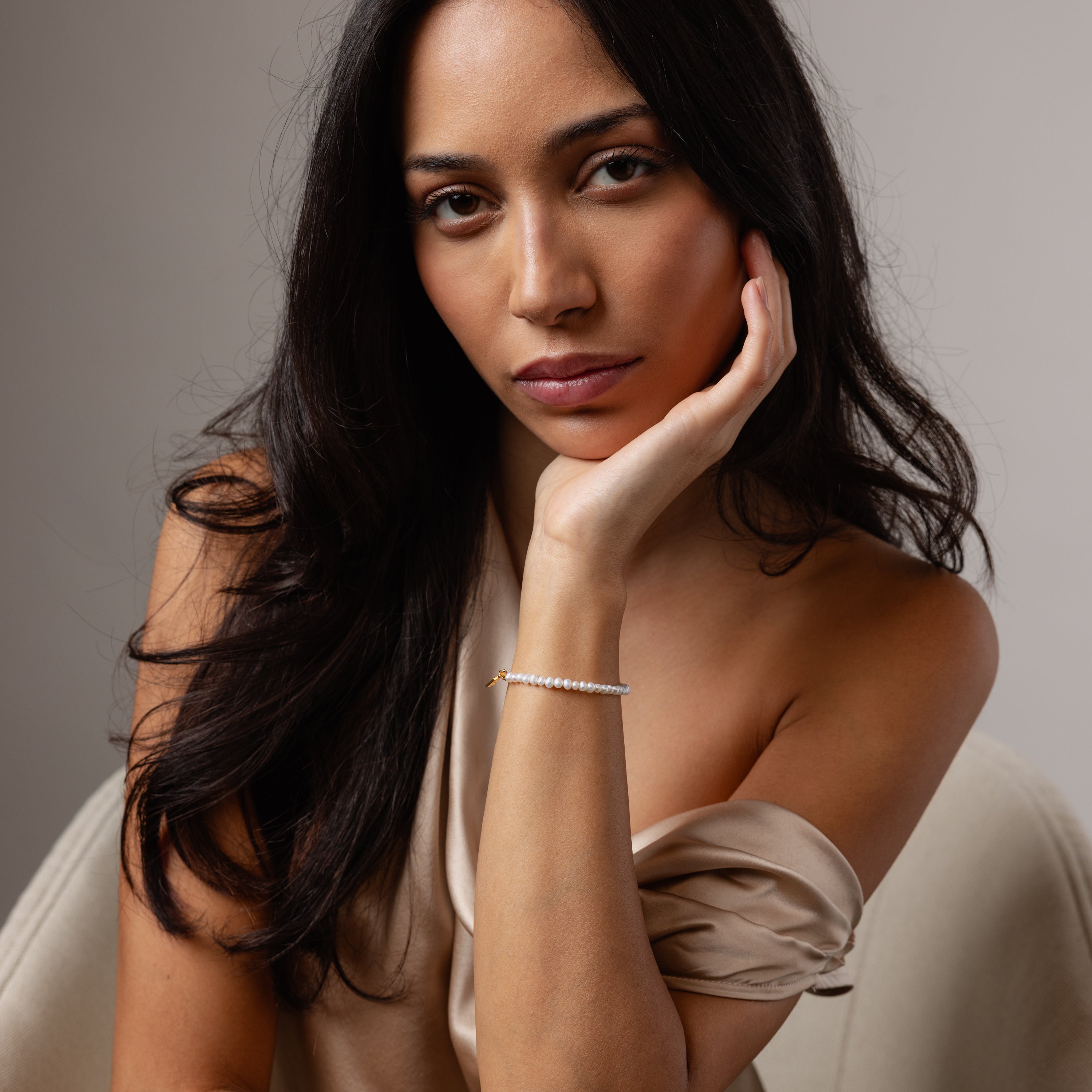 A woman with long dark hair, wearing an off-shoulder top, gazes thoughtfully while resting her face on her hand, adorned with elegant jewelry including the delicate Venus Pearl Bracelet.