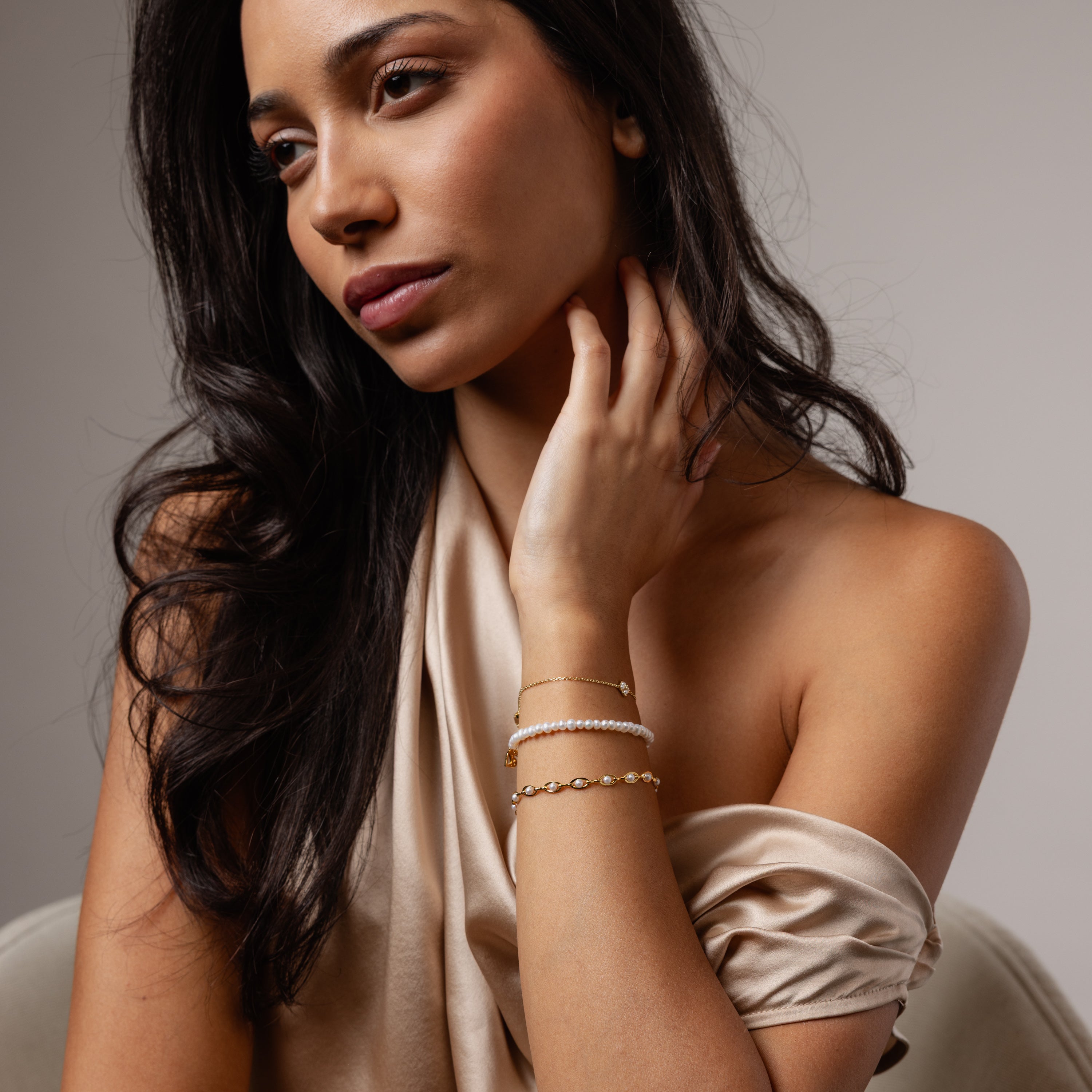 A woman in a silk off-shoulder dress wears elegant jewelry, including the Venus Pearl Bracelet, and gazes thoughtfully to the side.