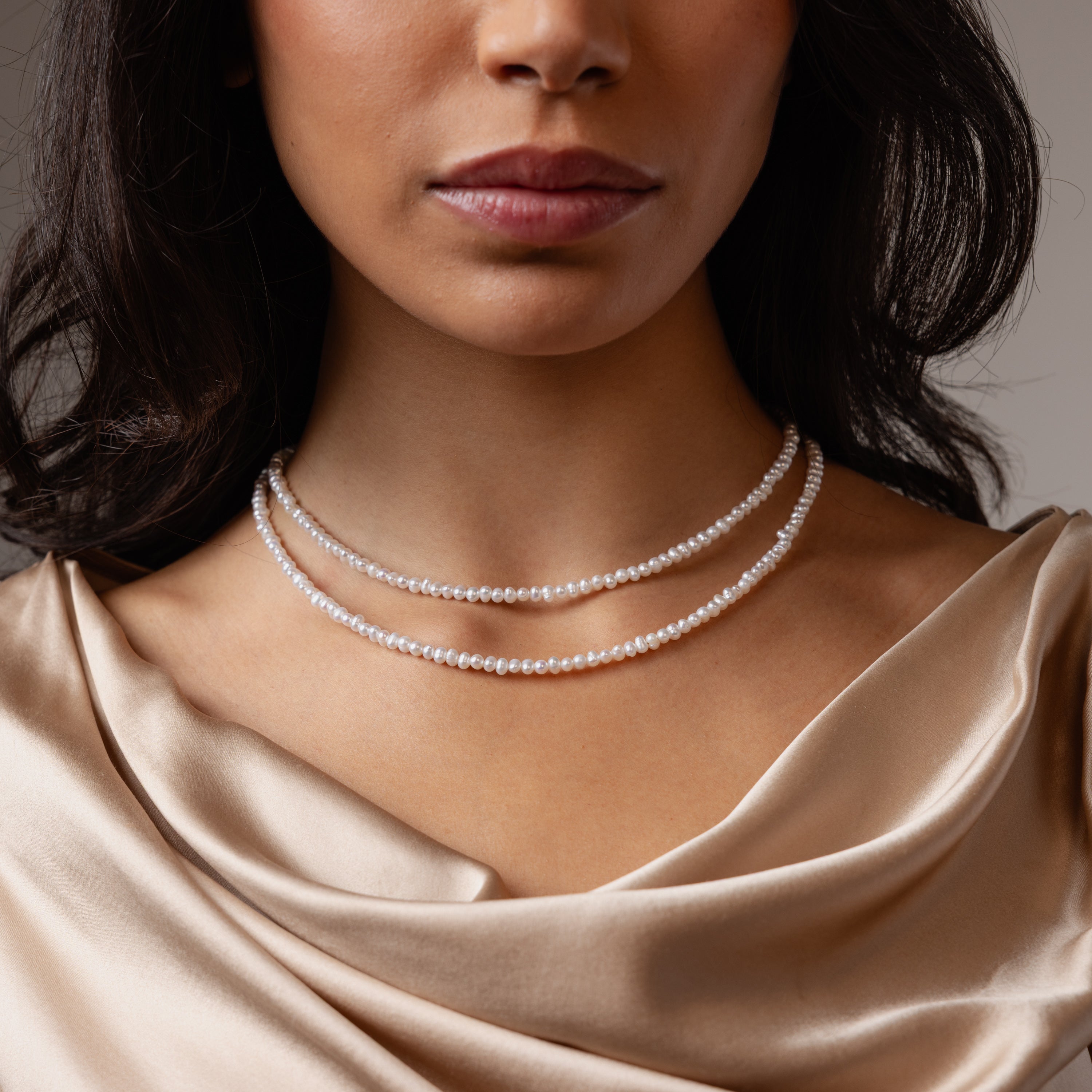 A woman wears the Venus Pearl Necklace—a double-strand freshwater pearl piece—paired with a silky, champagne-colored draped blouse for a timeless look.