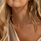 A woman with wavy blonde hair wears the Floating Pearl Necklace, featuring delicate freshwater pearls, paired with a silky beige top.